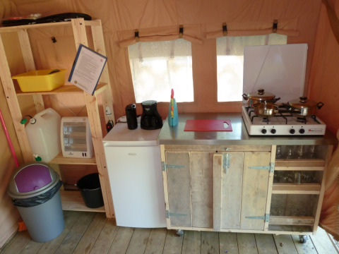 Pequeña cocina en una tienda safari con estufa, mini nevera, estantes, utensilios y papelera visible.