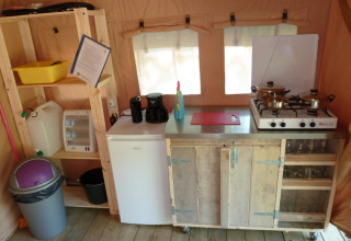 Pequeña cocina en una tienda safari con estufa, mini nevera, estantes, utensilios y papelera visible.