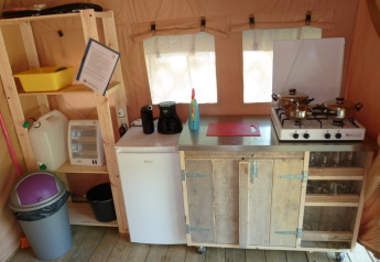 Small kitchen setup in a safari tent with stove, mini fridge, shelves, utensils, and trash bin visible.
