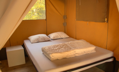 Interior de tienda safari con cama doble, dos almohadas y manta, iluminado por luz natural diurna.