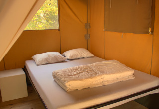 Interno di tenda safari con letto matrimoniale, due cuscini e una coperta illuminati da luce naturale.