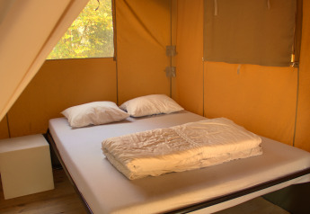 Interno di tenda safari con letto matrimoniale, due cuscini e una coperta illuminati da luce naturale.