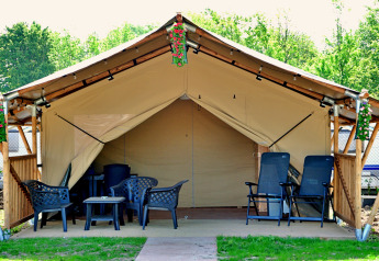 Safari tent met buitenstoelen, tafels en bloemen, omringd door groen en caravans op de achtergrond.