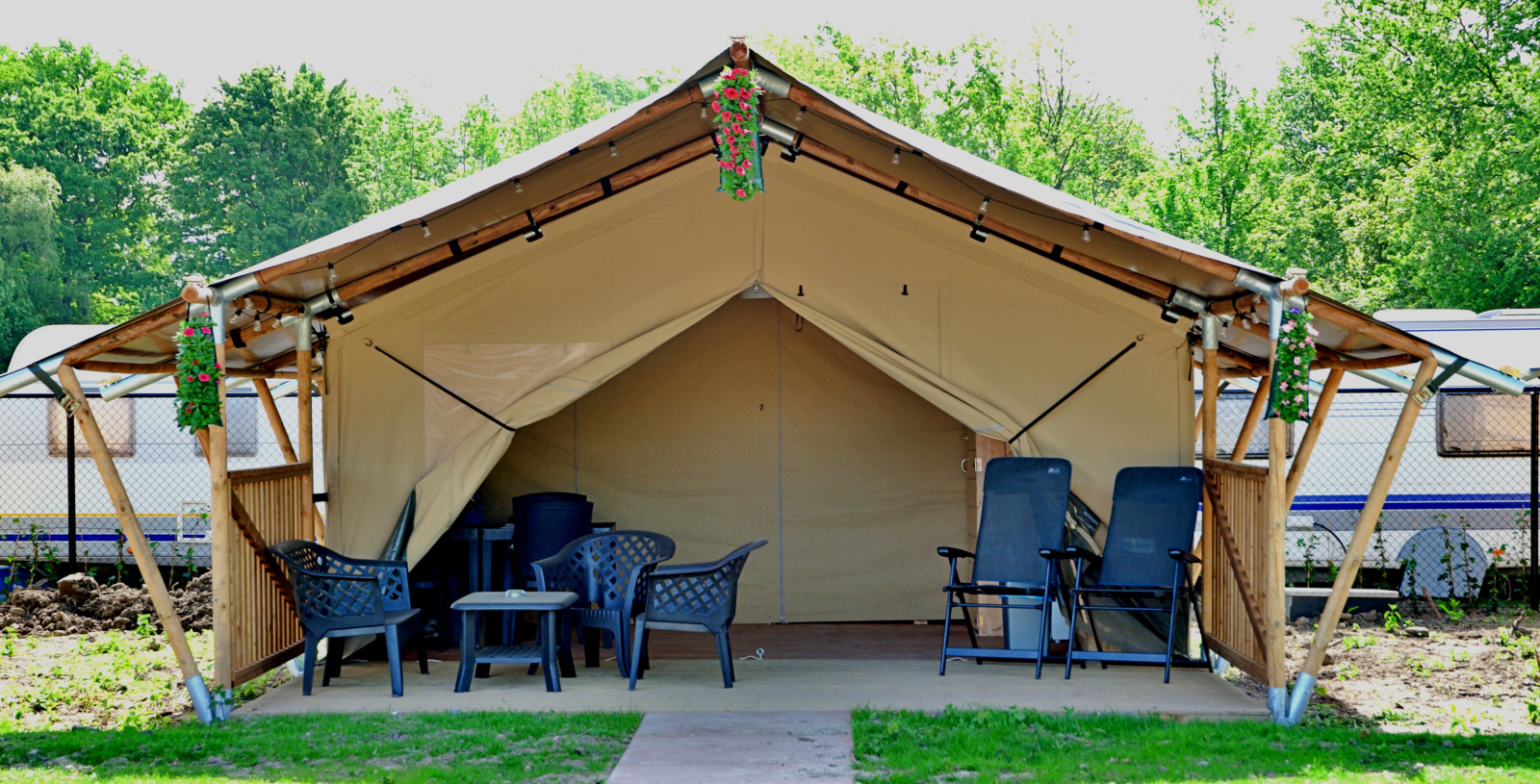 Safari tent met buitenstoelen, tafels en bloemen, omringd door groen en caravans op de achtergrond.