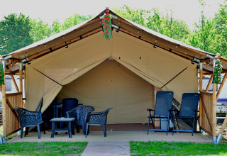 Safari tent met tuinmeubilair en bloemdecoratie, omgeven door groen en caravans op de achtergrond.