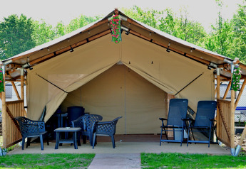 Safari tent met buitenstoelen, tafels en bloemen, omringd door groen en caravans op de achtergrond.