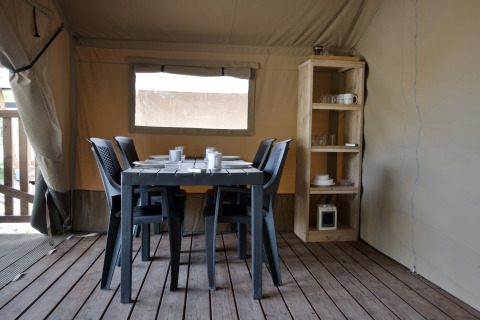 Eethoek binnen een safaritent met zwarte tafel, zes stoelen en houten rek met servies.