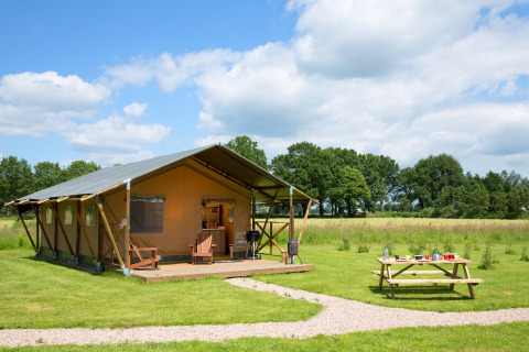 Une tente safari spacieuse appelée Lodgetent avec un porche en bois, située dans un champ herbeux avec une table de pique-nique à proximité.