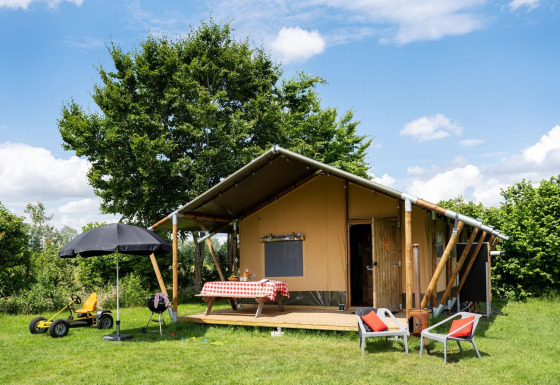 Buitenbeeld van een safaritent met veranda, geruite tafel, stoelen en speelgoed op een grasveld.