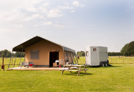 Tenda safari su prato verde con rimorchio, tavolo da picnic e paesaggio rurale alle spalle.