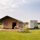 Tienda tipo safari en un campo verde junto a un remolque, mesa de pícnic y paisaje campestre de fondo.
