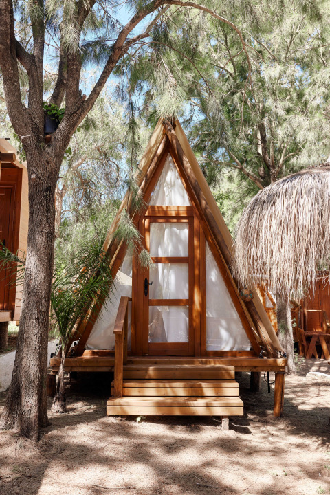 A-frame wooden cabin called Breña Tent at TAIGA Tarifa in Spain, nestled among trees in dappled sunlight.