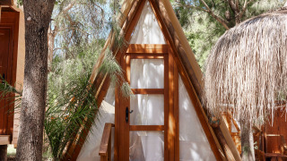 A-frame wooden cabin called Breña Tent at TAIGA Tarifa in Spain, nestled among trees in dappled sunlight.