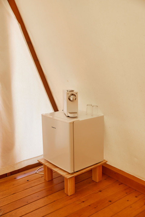 A small fridge on a wooden floor with two glasses and a coffee maker on top, inside a glamping tent.