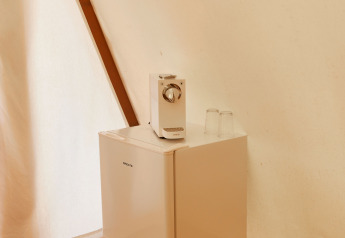 A small fridge on a wooden floor with two glasses and a coffee maker on top, inside a glamping tent.
