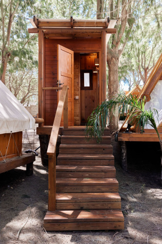 Cabaña de madera en un alojamiento glamping, con escalera y puerta abierta, rodeada de árboles.
