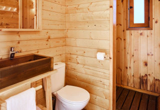 Salle de bain d’un hébergement glamping avec murs en bois, toilettes, lavabo en bois et petite fenêtre.