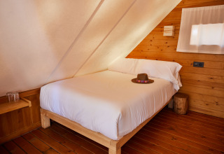 Bed met witte lakens en een hoed in Tamaral Tent bij TAIGA Tarifa, Spanje, met houten muren en vloer.