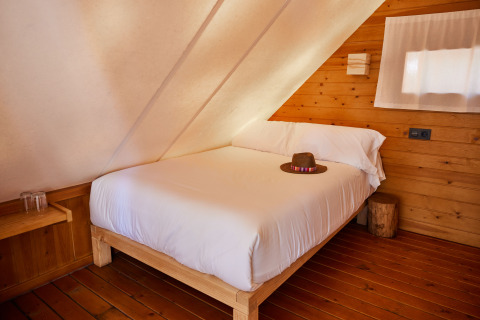 Bed met witte lakens en een hoed in Tamaral Tent bij TAIGA Tarifa, Spanje, met houten muren en vloer.