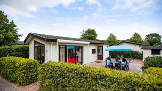 Casa de vacaciones con terraza y sombrilla azul en EuroParcs Noordwijkse Duinen, Países Bajos, tres personas afuera.