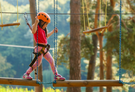Niña con casco y arnés cruza un circuito de cuerdas en un parque vacacional con alojamiento de glamping.