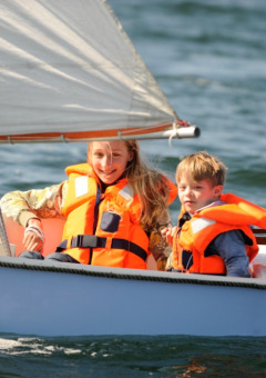 Deux enfants en gilets de sauvetage naviguent en petit voilier dans un parc de vacances proposant du glamping.