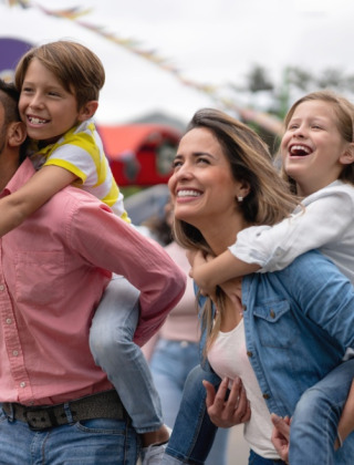 En glad familie nyder udendørs aktiviteter i en feriepark med glamping, børn får rygpiggy af forældrene.