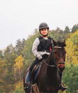 Niño con casco y chaleco monta a caballo en un parque vacacional con glamping, bosque de fondo.