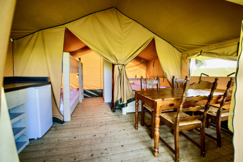 Interior view of a safari tent with a wooden dining table, bunk beds, refrigerator, and wood floor.