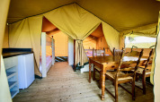 Interior view of a safari tent with a wooden dining table, bunk beds, refrigerator, and wood floor.