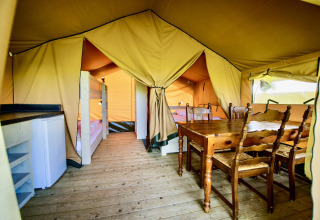 Interior view of a safari tent with a wooden dining table, bunk beds, refrigerator, and wood floor.