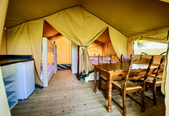 Vue intérieure d'une tente safari avec table à manger en bois, lits superposés, réfrigérateur et plancher.