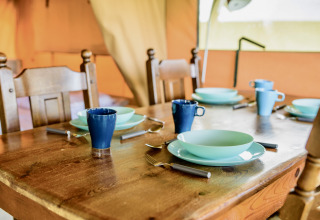 Mesa de madera puesta en una tienda safari con vajilla y tazas azules, preparada para cuatro personas.