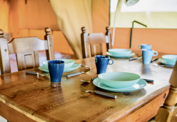 Mesa de madera puesta en una tienda safari con vajilla y tazas azules, preparada para cuatro personas.