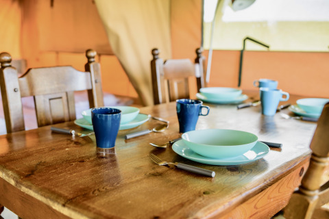 Table en bois dressée dans une tente safari avec vaisselle bleue et mugs, prête pour quatre personnes.