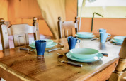 Mesa de madera puesta en una tienda safari con vajilla y tazas azules, preparada para cuatro personas.