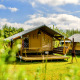 Tente safari moderne avec terrasse en bois par beau temps, sur un terrain de camping verdoyant et ensoleillé.