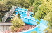 Familien genießen eine blaue Wasserrutsche im Veluwepark de Bosgraaf, umgeben von grünen Bäumen.