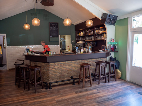 Zona de bar acogedora en Recreatiepark de Wrange, Gelderland, Países Bajos, con taburetes de madera y camarera.