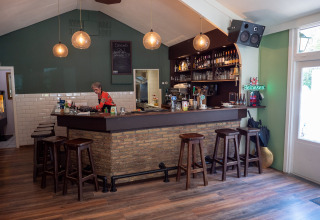 Zona de bar acogedora en Recreatiepark de Wrange, Gelderland, Países Bajos, con taburetes de madera y camarera.