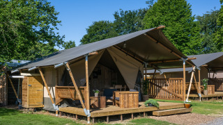 Safari-telt med sanitære faciliteter, træveranda og møbler, omgivet af træer under blå himmel.