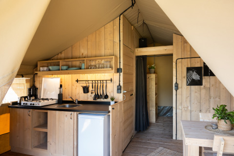 Vista interna di una tenda safari con pareti in legno, angolo cottura, zona pranzo e bagno privato.