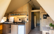 Vista interna di una tenda safari con pareti in legno, angolo cottura, zona pranzo e bagno privato.