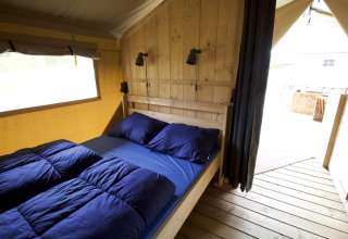 Binnenruimte van een safaritent op Camping Mareveld, houten wand, dubbel bed met blauw linnengoed en open deur.