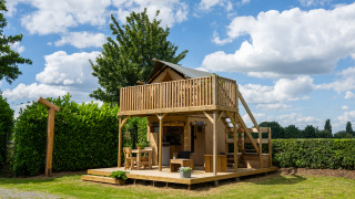 Tenda safari con servizi igienici, terrazza in legno e balcone, circondata da siepi e alberi verdi.