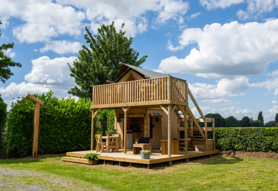 Tenda safari con servizi igienici, terrazza in legno e balcone, circondata da siepi e alberi verdi.