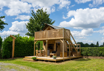 Tienda safari con instalaciones sanitarias, terraza de madera y balcón, rodeada de setos y árboles verdes.