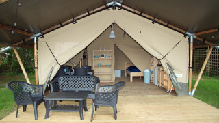 Tenda safari con piattaforma in legno, mobili da giardino neri e interni visibili con letti ed estagere.