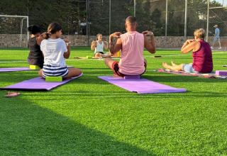 Folk deltager i en udendørs yogatime på grøn plæne i Camping del Mar, Catalonien, Spanien, solrig dag.