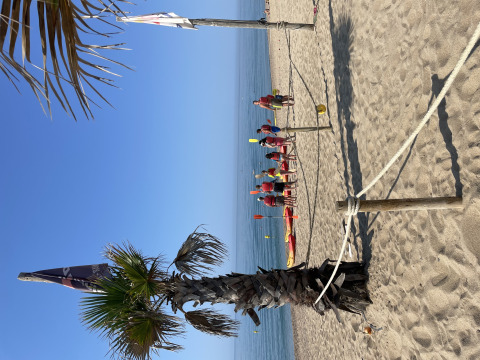 Escena de playa en Camping del Mar, Cataluña, España, con palmera, kayaks y gente con equipos rojos.